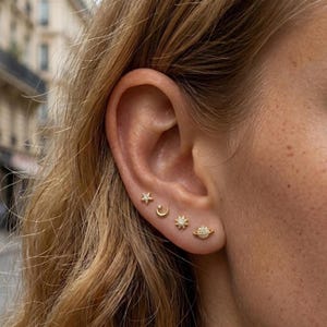 May include: A set of four gold-toned stud earrings worn in a single earlobe. The earrings feature celestial designs including a small star, a crescent moon, a sun with rays, and a planet with a ring, all accented with tiny clear stones.