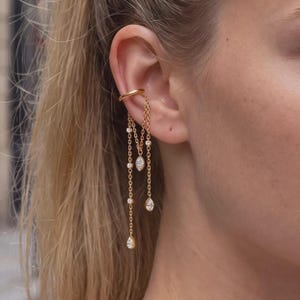 Può includere: Un ear cuff in oro con due catenelle delicate che pendono verso il basso. Le catene sono decorate con piccole pietre chiare e scintillanti a forma di marquise e goccia, creando un effetto a strati lungo il lato dell'orecchio.