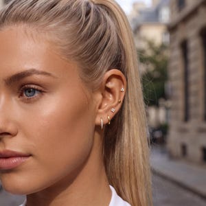 May include: A set of gold-toned earrings worn in a single ear, featuring two small hoop earrings, a flower-shaped stud with clear stones, a plain gold ball stud, and a three-stone cluster stud, all arranged along the earlobe and cartilage.