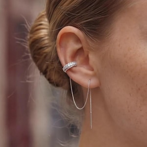 May include: A silver-toned ear cuff featuring a row of small, clear, sparkling stones along the outer band. A delicate, thin silver chain connects the cuff to a straight, slender metal post that hangs vertically from the earlobe.