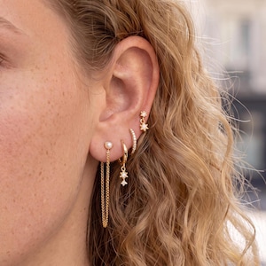 May include: A set of four gold-toned earrings worn in one ear. The jewelry includes a small star-shaped stud, a thin pavé hoop, a small hoop with a dangling star charm, and a stud with a dangling chain connected to a small pearl.