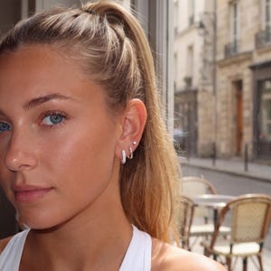 May include: A set of three silver-toned earrings worn in one ear, consisting of a small plain hoop, a slightly larger hoop featuring a row of clear stones, and a tiny stud earring with a single clear stone, all displayed on a side profile.