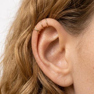 May include: A set of three thin gold ear cuffs worn on the upper cartilage. The bottom cuff features a delicate crisscross design, while the two cuffs positioned above it are simple, smooth, circular bands that hug the edge of the ear.