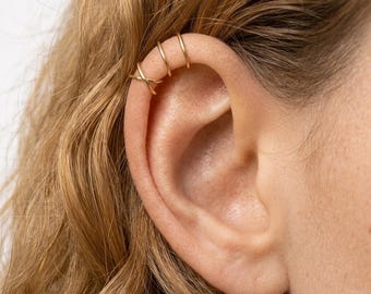 Puede incluir: Un conjunto de tres finos pendientes de tipo ear cuff dorados colocados en el cartílago superior. El pendiente inferior presenta un delicado diseño entrecruzado, mientras que los dos superiores son aros circulares lisos y sencillos.