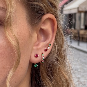 Pode incluir: Um conjunto de brincos dourados composto por uma pequena argola com pedras vermelhas e verdes alternadas, um brinco de pino vermelho e um brinco ear climber curvo com pedras vermelhas, brancas e verdes cravadas.