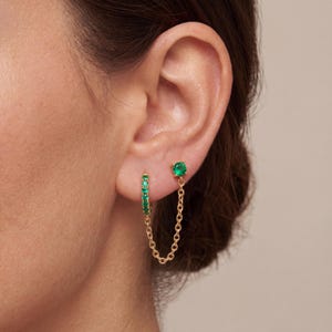 May include: A gold-toned hoop earring featuring a row of small green stones, connected by a delicate gold chain to a single green stone stud earring. The jewelry is worn in two piercings on the earlobe, creating a linked, layered appearance.