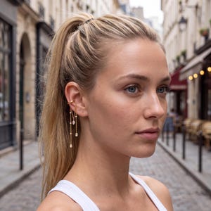 May include: A gold ear cuff featuring two delicate chains that drape downward, each adorned with small, clear, teardrop-shaped stones. The jewelry piece secures to the upper cartilage of the ear, creating a layered, dangling effect with metallic links.