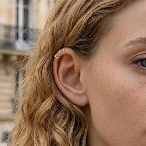 May include: A gold ear cuff featuring a delicate design with three thin, parallel bands that converge into a single point at the top, worn on the upper cartilage of the ear. The jewellery has a polished metallic finish and a simple, slender structure.
