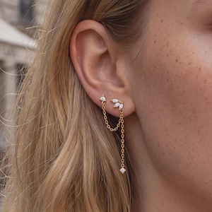 Peut inclure: Une boucle d'oreille double en métal doré composée d'un petit clou avec une pierre transparente et d'un autre clou orné de trois pierres taille marquise en forme de feuille, reliés par une fine chaîne dorée avec une pierre pendante.