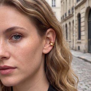 May include: A gold-toned ear cuff featuring a double-band design with a smooth, polished finish. The jewelry piece consists of two parallel, slightly curved bands that wrap around the outer edge of the ear cartilage, creating a layered appearance.
