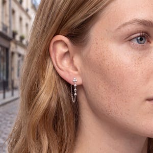 May include: A silver-toned earring featuring a vertical row of four small, clear, round-cut stones set in metal, with a delicate chain connecting the top and bottom of the stone setting to create a looped design that hangs from the earlobe.