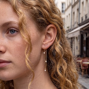 May include: A gold threader earring featuring a delicate chain design with three small, clear, round-cut stones spaced along the length. The earring hangs vertically from the earlobe, showcasing a minimalist metallic finish and sparkling accents.