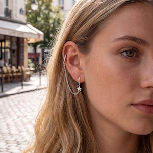 May include: A silver-toned ear cuff and hoop earring connected by a delicate chain. The ear cuff is a plain band, while the hoop features a small, sparkling star-shaped charm encrusted with tiny clear stones. The jewellery is worn on the ear.