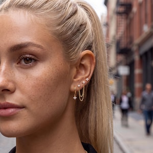 May include: A set of gold-toned earrings featuring a small hoop with a four-petaled flower charm, a single stud with a clear stone, and a double-chain earring connecting two four-petaled flower studs, all with clear stone accents.