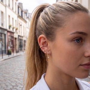 Könnte beinhalten: Eine goldene Ohrmanschette mit einer Reihe kleiner klarer Steine, ein goldener sternförmiger Ohrstecker und zwei goldene Creolen, die durch eine zarte goldene Kette mit einem kleinen Sternanhänger und einem zentralen klaren Stein verbunden sind.
