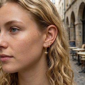 May include: A set of gold-toned earrings consisting of a small stud with a starburst design, a plain hoop, and a hoop featuring a dangling starburst charm. The jewellery is worn in three separate piercings along the earlobe and cartilage.