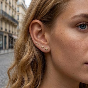May include: A set of four small gold stud earrings worn in a single earlobe. The earrings feature celestial designs including a tiny star, a crescent moon, a sunburst, and a planet with rings, all accented with small clear stones.