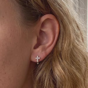 May include: A small silver-toned hoop earring featuring a cross-shaped charm adorned with clear, sparkling stones. The earring is secured in the earlobe, showcasing a metallic finish and a delicate, symmetrical design with multiple embedded crystals.