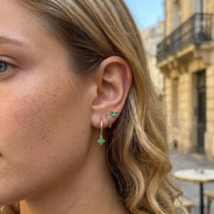 May include: A set of three gold earrings worn in one ear. The top earring is a small green stone stud, the middle is a small gold hoop with green stones, and the bottom is a gold hoop featuring a small, four-petaled green flower charm.