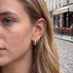 Könnte beinhalten: Drei goldfarbene Ohrringe an einem Ohrläppchen. Der größte Ohrring ist ein glatter, dicker Creolen-Ring. Der mittlere Ohrring ist ein kleinerer, dünnerer Ring mit strukturierter Oberfläche. Der kleinste Ohrring ist ein winziger, dünner Ring.