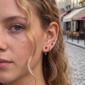 Puede incluir: Un conjunto de pendientes de oro con pequeños rubíes rojos y esmeraldas verdes de talla redonda. La joyería incluye un pendiente de botón, un aro pequeño y un pendiente trepador curvo, todos con piedras preciosas alternas en metal dorado.