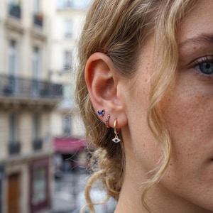 Op de afbeelding: Een set van drie goudkleurige oorbellen in één oor. De bovenste oorbel is een kleine blauwe vlinderknop, de middelste is een dunne ring met blauwe steentjes en de onderste is een gouden ring met een kleine bungelende bedel met een blauwe steen.