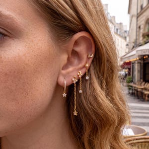 May include: A set of five gold-toned earrings featuring celestial motifs. The collection includes a small hoop with a star charm, two crescent moon and star drop earrings with delicate chains, a starburst stud, and a tiny crescent moon stud.