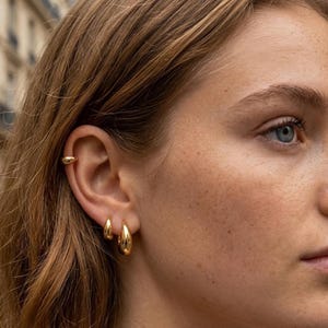 May include: A set of three gold-toned earrings consisting of one small smooth ear cuff positioned on the upper cartilage and two graduated teardrop-shaped hoop earrings worn in the earlobe, all featuring a polished reflective finish.