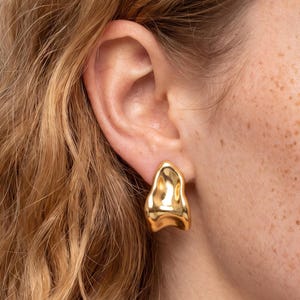 May include: A gold-toned earring with an irregular, organic, and molten-like shape. The surface is smooth and highly reflective, catching the light to create highlights and shadows across its curved, abstract form.