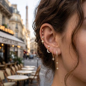 Könnte beinhalten: Nahaufnahme eines Ohrs, das mit mehreren goldenen Ohrringen geschmückt ist. Die Ohrringe umfassen einen Mond, einen Stern, einen Blitz und ein hängendes Design mit Sternen und kleinen Kristallen. Im Hintergrund eine Pariser Straßenszene.