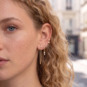 May include: A set of gold-toned earrings worn in the earlobe and cartilage. The jewellery includes a small star-shaped stud, a thin hoop with tiny clear stones, a small hoop with a dangling star charm, and a stud with a chain and pearl drop.