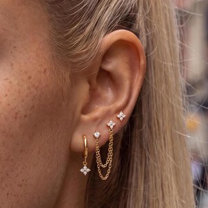 May include: A set of gold-toned earrings featuring a small hoop with a four-petaled flower charm, a single stud with a four-petaled flower, and a double-chain earring connecting a single stone stud to a four-petaled flower stud.