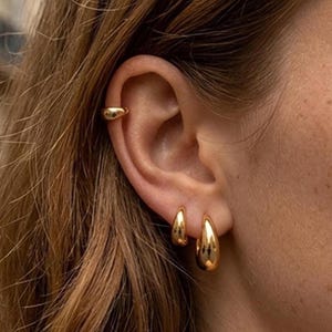Op de afbeelding: Een set van drie goudkleurige sieraden gedragen aan het oor, bestaande uit een gladde, gepolijste oorklem op het bovenste kraakbeen en twee bijpassende druppelvormige oorringen bevestigd aan de onderste oorlel.