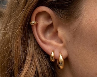 May include: A set of three gold-toned jewellery pieces worn on the ear, consisting of one smooth, polished ear cuff positioned on the upper cartilage and two matching teardrop-shaped hoop earrings attached to the lower earlobe.