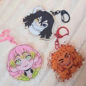 Könnte beinhalten: Drei Anime-Schlüsselanhänger aus Acryl mit Charakterporträts. Ein Schlüsselanhänger zeigt einen Charakter mit schwarzen Haaren und Bandagen, ein anderer hat rosa Haare und der dritte orangefarbene Haare. Jeder Schlüsselanhänger hat einen unterschiedlich farbigen Verschluss.