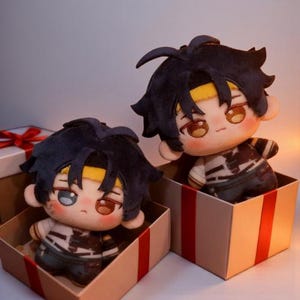 Puede incluir: Dos muñecos de peluche con pelo azul oscuro y cintas amarillas para la cabeza están sentados en cajas de regalo abiertas. Los muñecos tienen la piel clara, mejillas rosadas y visten trajes blancos y negros. Las cajas son beige con detalles de cinta roja.