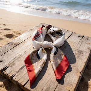 Peut inclure: Un foulard en soie crème avec des détails rouges, marine et dorés, noué sur une surface en bois vieilli. Le foulard présente un motif géométrique et équestre. L'arrière-plan montre la mer et une plage de sable.