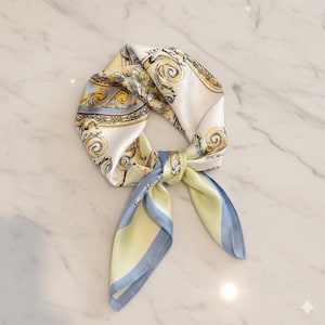 Peut inclure: Un foulard carré en soie avec un motif décoratif blanc, jaune et bleu. Le foulard est noué, avec les extrémités pendantes. Le foulard a une bordure bleu clair et jaune.