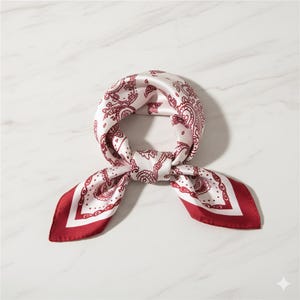 Op de afbeelding: Vierkante zijden sjaal met een wit en bordeaux paisley patroon. De sjaal is geknoopt en heeft effen bordeauxrode randen. Het paisley-ontwerp is bordeauxrood op een witte achtergrond.