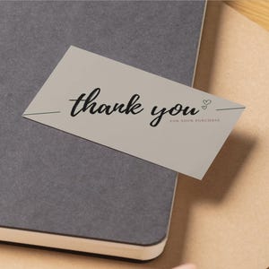 May include: A light gray thank you card with black script text and two small hearts. The card is on top of a gray notebook and a brown envelope. The text reads "thank you" and "for your purchase".