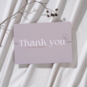 May include: A mauve-colored card with the words "Thank you" in a serif font, and a butterfly illustration. The text "FOR YOUR PURCHASE" is below the main text. A twig with small berries and a bare branch are in the background.