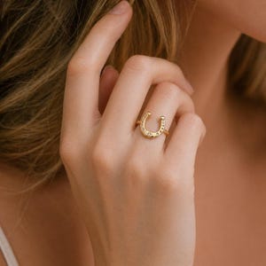 May include: A gold-toned ring featuring a horseshoe-shaped centerpiece. The horseshoe design is embellished with small, clear, round-cut stones set along its curve, with a single, slightly larger, star-shaped accent at the base of the horseshoe.