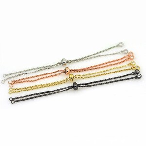 May include: An assortment of adjustable chain bracelets in silver, copper, gold, and black. Each bracelet features a sliding bead for size adjustment and is adorned with small, clear gemstones at the ends. The bracelets are displayed on a white background.