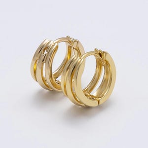 May include: A pair of gold-colored hoop earrings. Each earring features a triple-layered design, creating a bold and textured appearance. The earrings have a polished, shiny finish, reflecting light. The hoops are set against a plain white background.