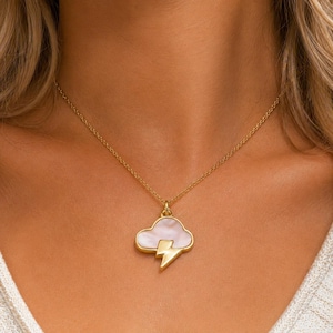 May include: A gold-toned necklace featuring a pendant shaped like a cloud with a lightning bolt. The cloud portion is filled with a smooth, iridescent white material, while the lightning bolt and the surrounding frame are crafted in gold metal.