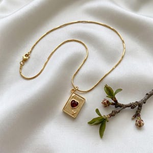 May include: A gold-toned necklace featuring a rectangular pendant shaped like a playing card. The pendant displays the letter A at the top and bottom corners with a central, raised red heart emblem. The chain is a thin, flexible gold-toned metal.