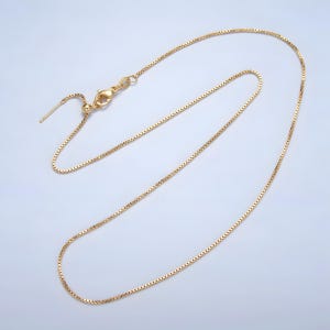 May include: A gold-colored box chain necklace with an adjustable clasp. The chain is made of small, square links and has a delicate appearance. The necklace is displayed on a light background.