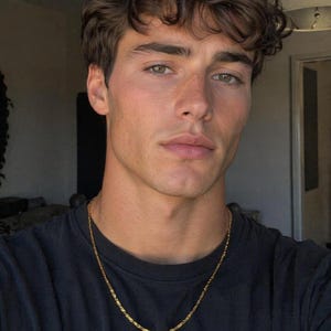 May include: A portrait of a person with brown curly hair and green eyes. They are wearing a black t-shirt and a gold chain necklace. The background is blurred, with a focus on the person's face.