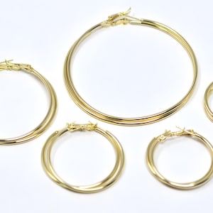 May include: Five gold-colored hoop earrings of varying sizes are arranged on a white surface. The earrings have a classic, circular design with a polished finish. The largest hoop has a diameter of approximately 3 inches.