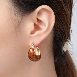 May include: Gold-tone hoop earrings with a brown, glossy, teardrop-shaped design. The earrings are worn on an ear, showcasing the contrast between the gold and the rich brown color. The earrings have a modern, elegant style.